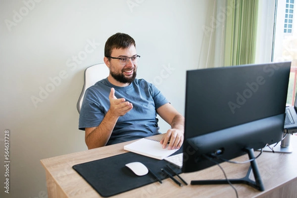 Fototapeta Handsome man smiling with a beard and glasses of сaucasian talks on a video call with colleagues at the workplace with monitors. Programmer, manager, economist, freelancer, having video chat, waving