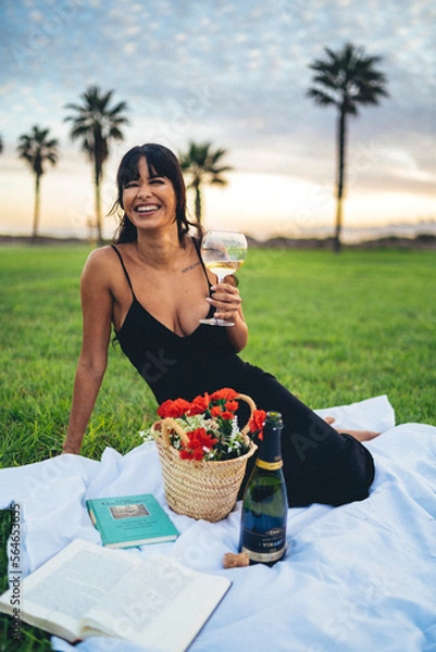 Obraz Chica morena guapa de picnic leyendo un libro y tomando vino con flores en un cesped cerca de la playa