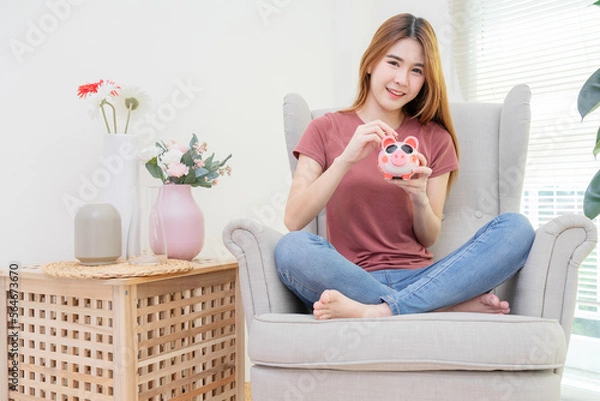 Obraz Young woman very happy putting coin into piggy bank for saving money on sofa near window and flower vase