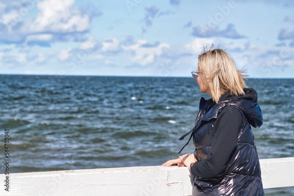 Fototapeta A blond-haired woman looks straight ahead into the endless sea. She has glasses and is wearing a black sweatshirt and a black warmer. She is standing with her hands on the railing of the pier.
					