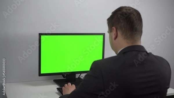 Fototapeta A young businessman is sitting in front of a computer with a green screen. A man in a suit works for a PC. Screen with chroma key.