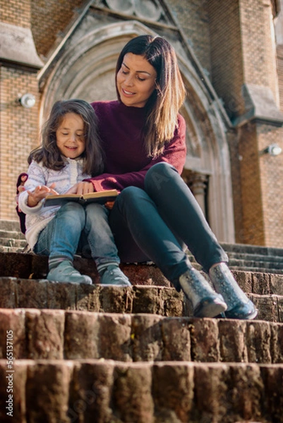 Fototapeta Mother and daughter sitting at the stairs and read a book outdoor. Low angle view. 