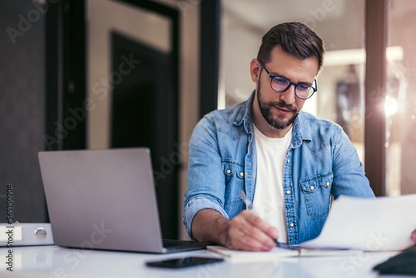 Obraz Satisfied young man with glasses sitting at a desk and doing paperwork at his workplace.
