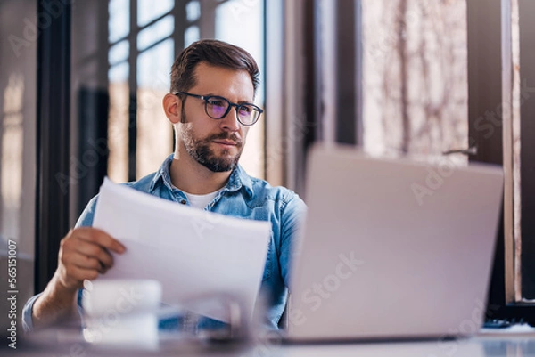 Obraz Satisfied young man with glasses sitting at a desk and doing paperwork at his workplace.