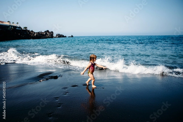 Fototapeta Cute little girl wearing in bright colorful swimsuit. standing and playing on black sand beach on Tenerife near Atlantic ocean. funny summer time, summer vacation destination