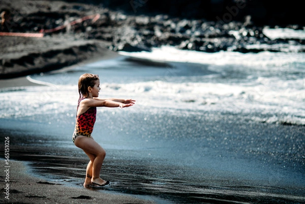 Fototapeta Cute little girl wearing in bright colorful swimsuit. standing and playing on black sand beach on Tenerife near Atlantic ocean. funny summer time, summer vacation destination