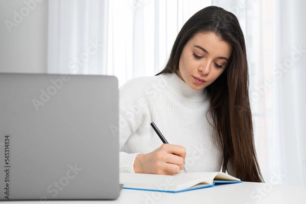 Obraz Young woman takes online courses via notebook making notes while listening to teacher. Attentive student sits at table in room studying online at home