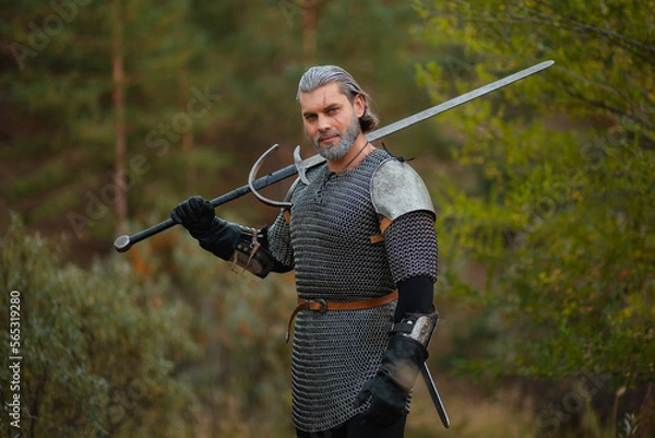 Fototapeta An aged male warrior in chain mail with gray hair and a scar on his face. Knight in armor holds a two-handed sword.