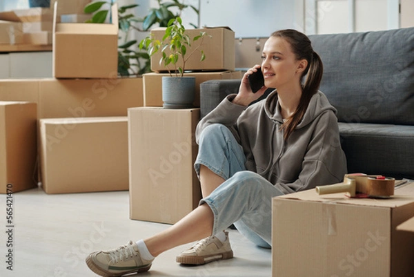 Fototapeta Young woman discussing her move with loaders by the mobile phone while sitting on the floor among packed boxes