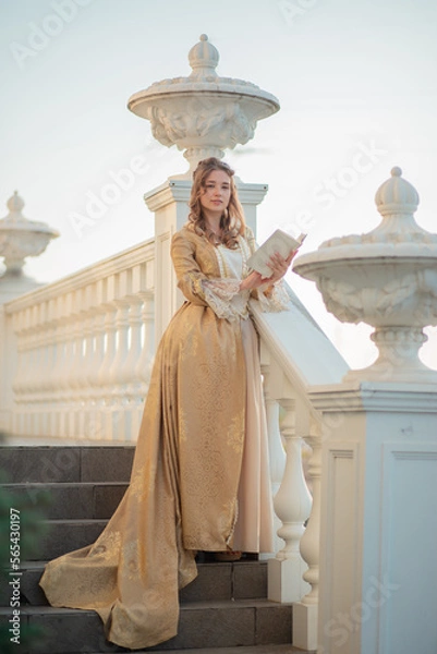 Fototapeta A beautiful young woman in an eighteenth century historical gold dress stands on the stairs of a mansion with a book in her hands. the princess is reading in the palace.