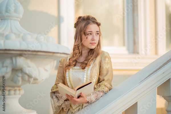 Fototapeta A beautiful young woman in an eighteenth century historical gold dress stands on the stairs of a mansion with a book in her hands. the princess is reading in the palace.