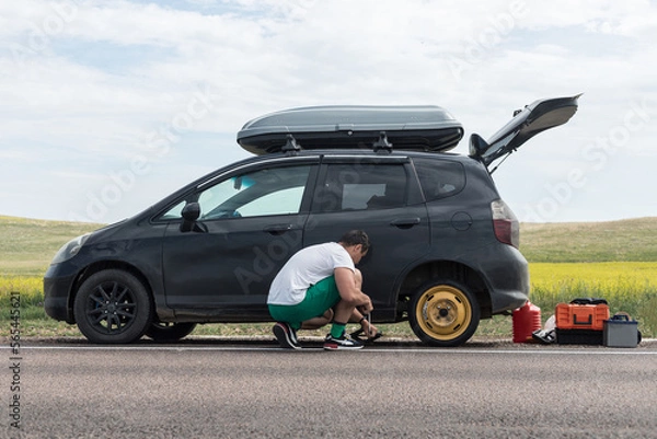 Fototapeta A man lifts a car to replace a tire after a puncture on the side of the road using a hydraulic jack inserted under the body, lifts the car and puts a spare wheel