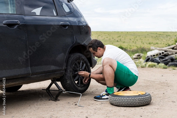 Fototapeta Young man changing the punctured tyre on his car loosening the nuts with a wheel spanner before jacking up the vehicle