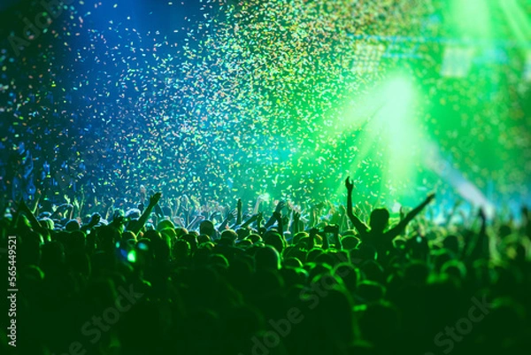 Fototapeta A crowded concert hall with scene stage green lights, rock show performance, with people silhouette, colourful confetti explosion fired on dance floor air during a concert festival