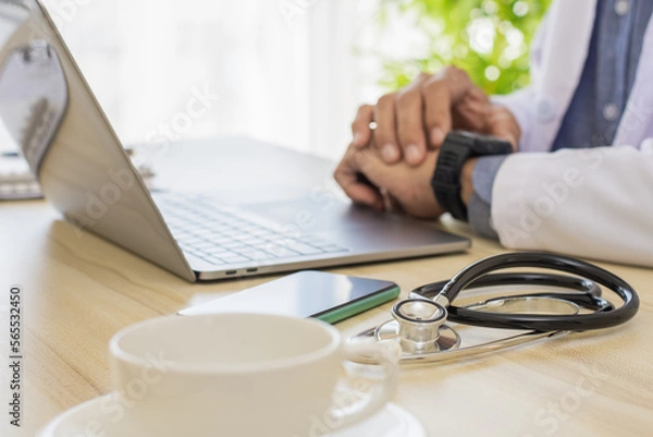 Obraz Male doctor looking at smart watch while work on laptop computer with medical stethoscope and mobile smartphone on the desk at clinic or hospital.