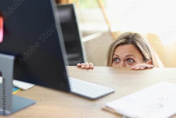 Fototapeta Scared female manager hidden behind office desk and looking at computer screen.
