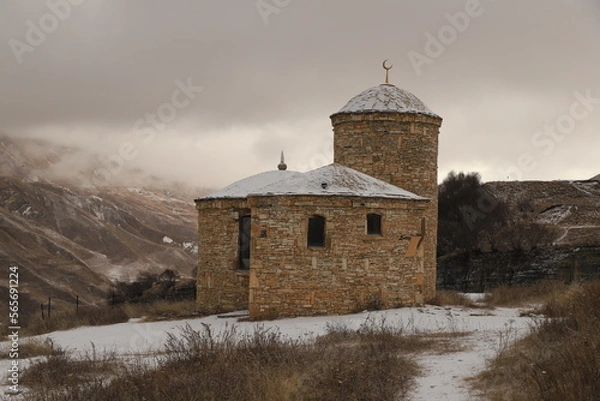 Fototapeta An ancient stone Muslim temple in the mountains of Dagestan - stands on a snowy slope against the backdrop of mountains and a gloomy sky.