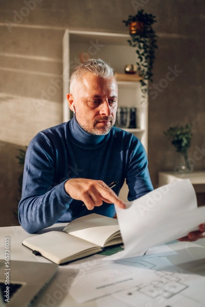 Fototapeta Middle aged man working in a home office and reading paper documents