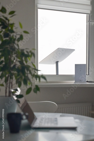 Fototapeta KYIV, UKRAINE - JANUARY 24, 2023: Starlink antenna and wi-fi router on windowsill in home office interior with laptop on table with cup of coffee in foreground, selective focus.