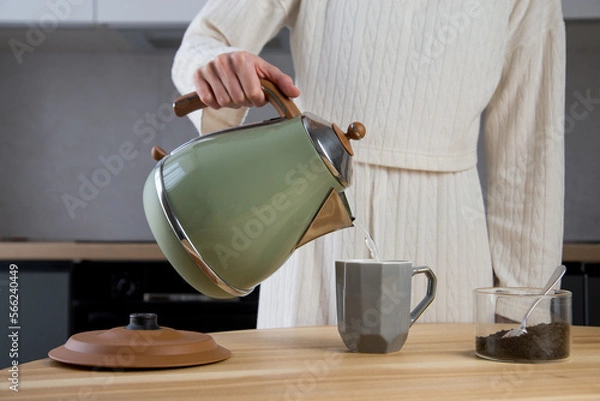 Fototapeta woman prepares tea in the kitchen on a wooden table. adding tea leaves to a cup and pouring hot water with an electric kettle