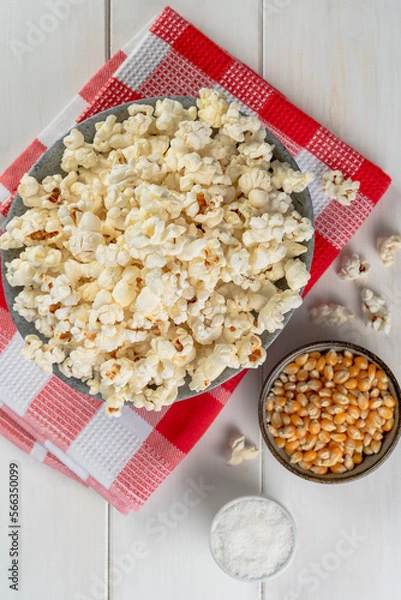 Obraz bowl with popcorn, popcorn kernels and salt on a red checkered napkin, on a white wooden table, top view.