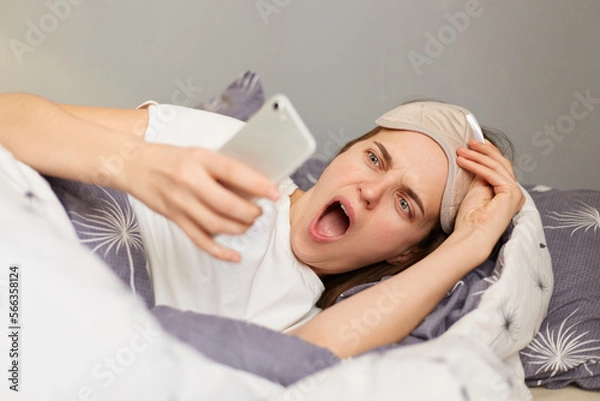 Fototapeta woman in bed with a sleep mask wakes up and talks on the phone. an unexpected call on a smartphone early in the morning or at night. a tired, sleepy European woman answers work calls. insomnia