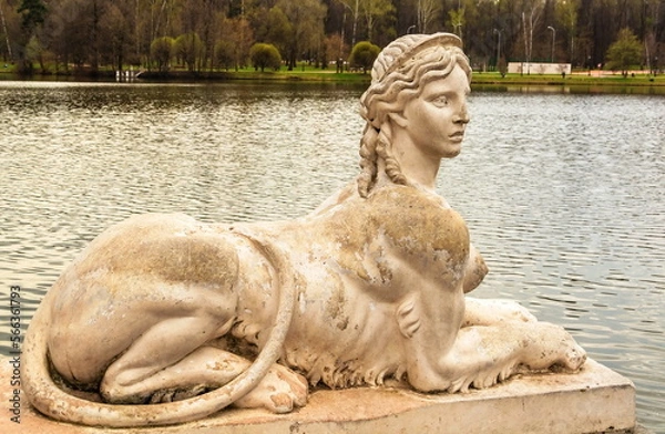 Fototapeta An ancient park sculpture in the form of a female lion sphinx on the shore of a pond in the park
