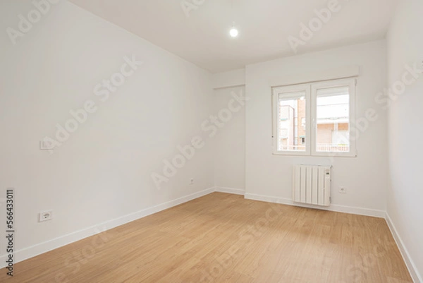 Fototapeta Empty living room with laminated flooring and recently finished smooth white walls with an aluminum radiator under a window of the same material