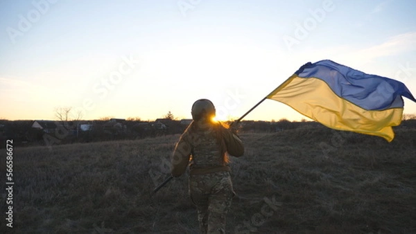 Obraz Female ukrainian army soldier runs and waves flag of Ukraine at sunset. Woman in military uniform lifted up flag in honor of the victory against russian aggression. Invasion resistance concept. Slowmo