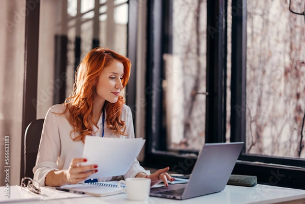Obraz Happy smiling businesswoman in formal wear sitting at desk in modern office in front of laptop and doing paperwork