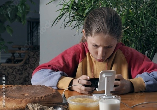 Fototapeta A teenager boy sits at the dinner table, ignoring food, uses the phone for computer games in social networks. Hobbies of modern children. Use of modern technologies by the younger generation