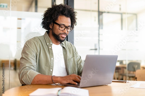 Fototapeta Focused curly handsome man using laptop computer in contemporary coworking space, freelance programmer in glasses coding sitting at the office desk, creativity designer or architect typing