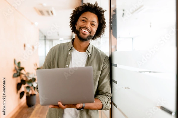 Obraz Inspired young african-american businessman looks at camera and smiles cheerfully, standing in modern office and using laptop. Male office employee, freelancer, designer full of new ideas and creative