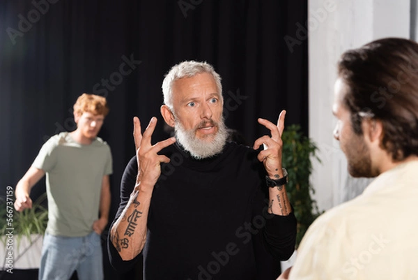 Fototapeta bearded art director gesturing while talking to blurred actor in theater