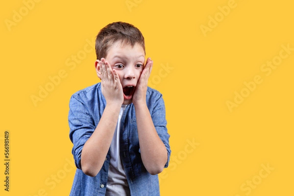 Fototapeta A portrait of a schoolboy showing emotion of shock. He’s holding his cheeks and screaming. He’s looking right and down. Yellow background. Front view. Copy space