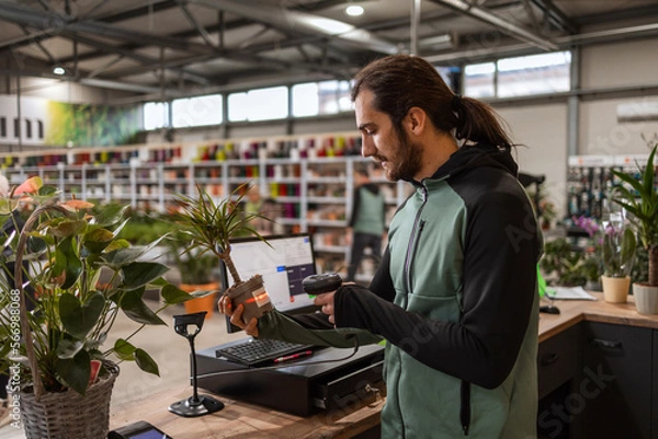 Fototapeta A man behind the cashier stand is scanning a price tag on a potted plant with a barcode reader.