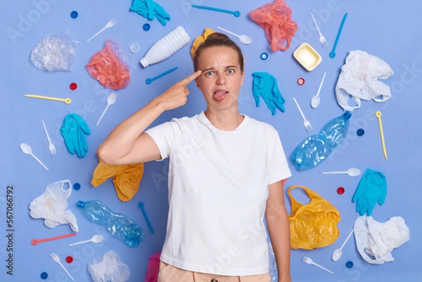 Obraz Portrait of funny woman dressed in white T-shirt surrounded with picked plastic litter isolated on blue wall, making suicide gesture and showing tongue out, being tired of pollution.