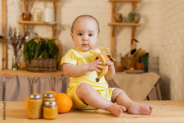 Fototapeta a cute kid in a yellow bodysuit is sitting in the kitchen with cans of baby food. The first complementary feeding of the child. Baby purees