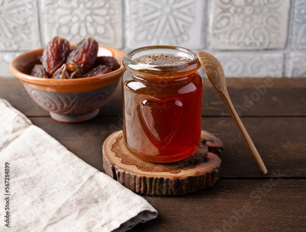 Obraz Natural organic healthy date syrup in a glass jar with whole dates and wood spoon on the background