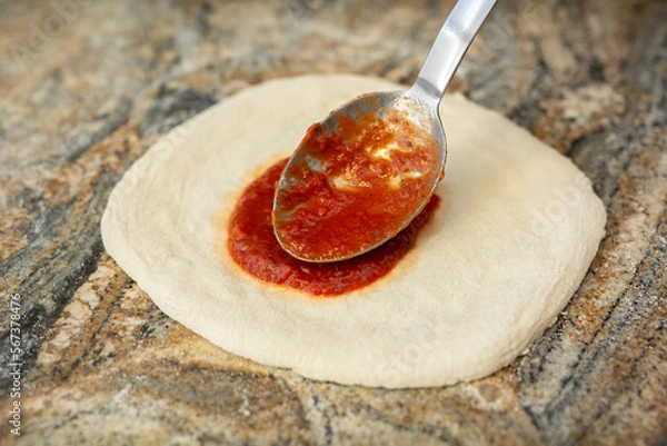 Fototapeta A man adding tomato sauce to the base of an Italian pizza. Cooking pizza in a pizzeria.