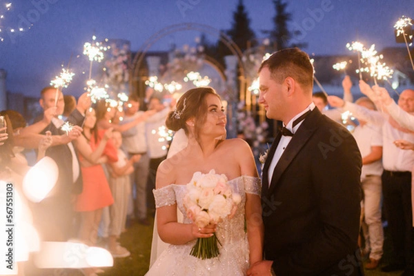 Obraz Close view of happy newlyweds in elegant dress and suit, looking and smiling to each other on background of arch, which created by guests with sparkles during evening ceremony