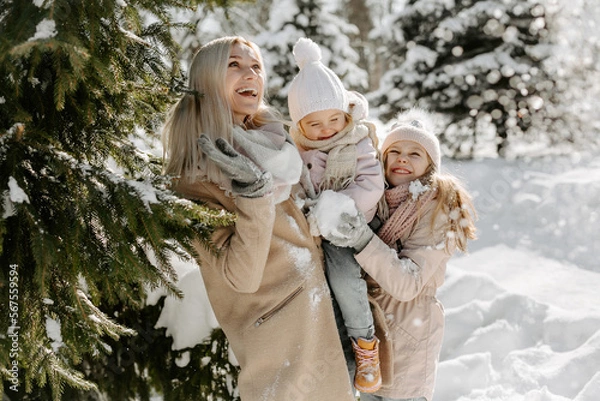 Fototapeta Front view of mother with daughters enjoying winter day in forest. Loving mother standing near green tree with daughters and playing with snow during winter vacation