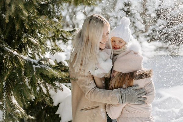 Fototapeta Portrait of mother spending time with children during winter holiday in snowy park. Smiling woman, dressed in warm outwear, embracing her daughters, while standing near tree during light snow falling