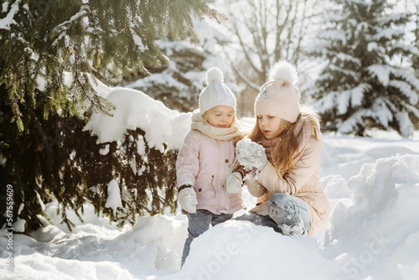 Fototapeta Front view of happy children sculpting snowman in a snowy park. Older sister, holding snowball and playing with little sister while standing at sunny winter park