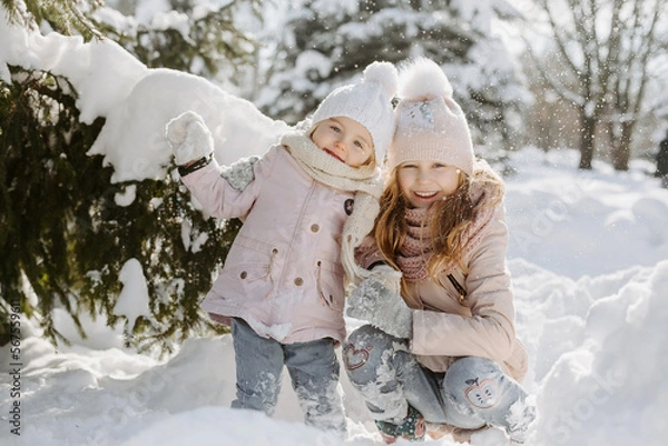 Fototapeta Portrait of cute little girls posing in snowy forest. Happy sisters, wearing warm clothes and scarves, holding with hands and standing in background of sunny snowy park