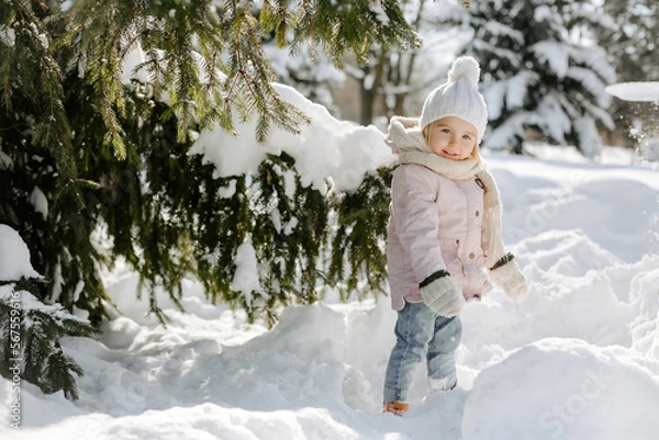 Fototapeta Portrait of a cute little girl posing alone in a snowy park. Happy child, standing near snowy twig of Christmas tree and looking at camera