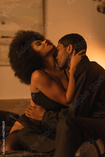 Fototapeta side view of african american man hugging and kissing seductive girlfriend in bedroom at night