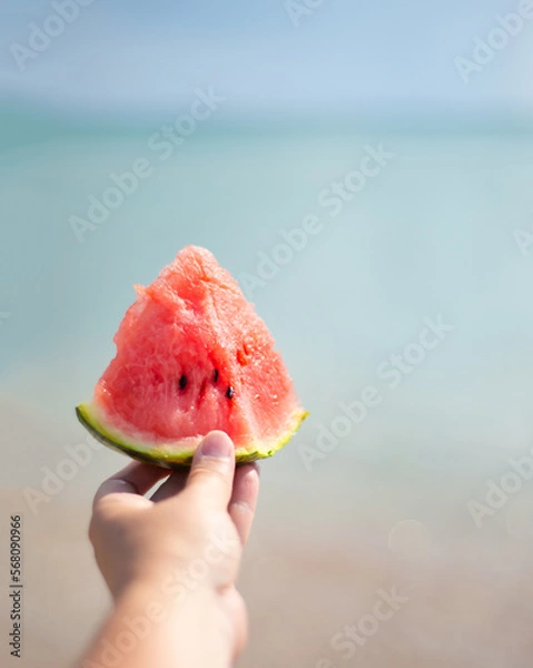 Fototapeta Watermelon slice in hand. Beach and sea. Ocean coast. Picnic on the coast. Vegetarian food in nature