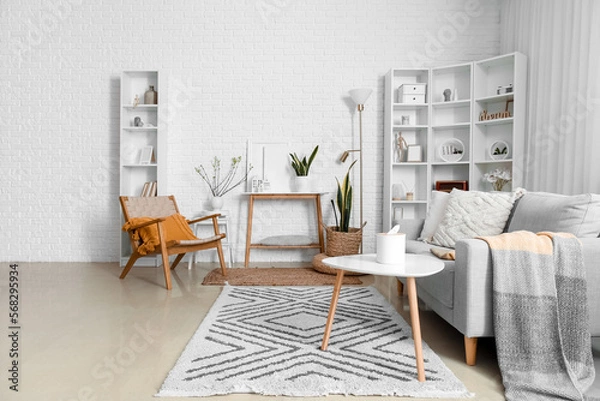 Fototapeta Interior of living room with armchair, sofa and house candle holders on table near white brick wall