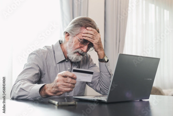 Fototapeta Upset senior elderly man holding credit card by laptop having trouble worry finance safety data or online payment security. Bank client concerned about problem with credit card, financial fraud threat